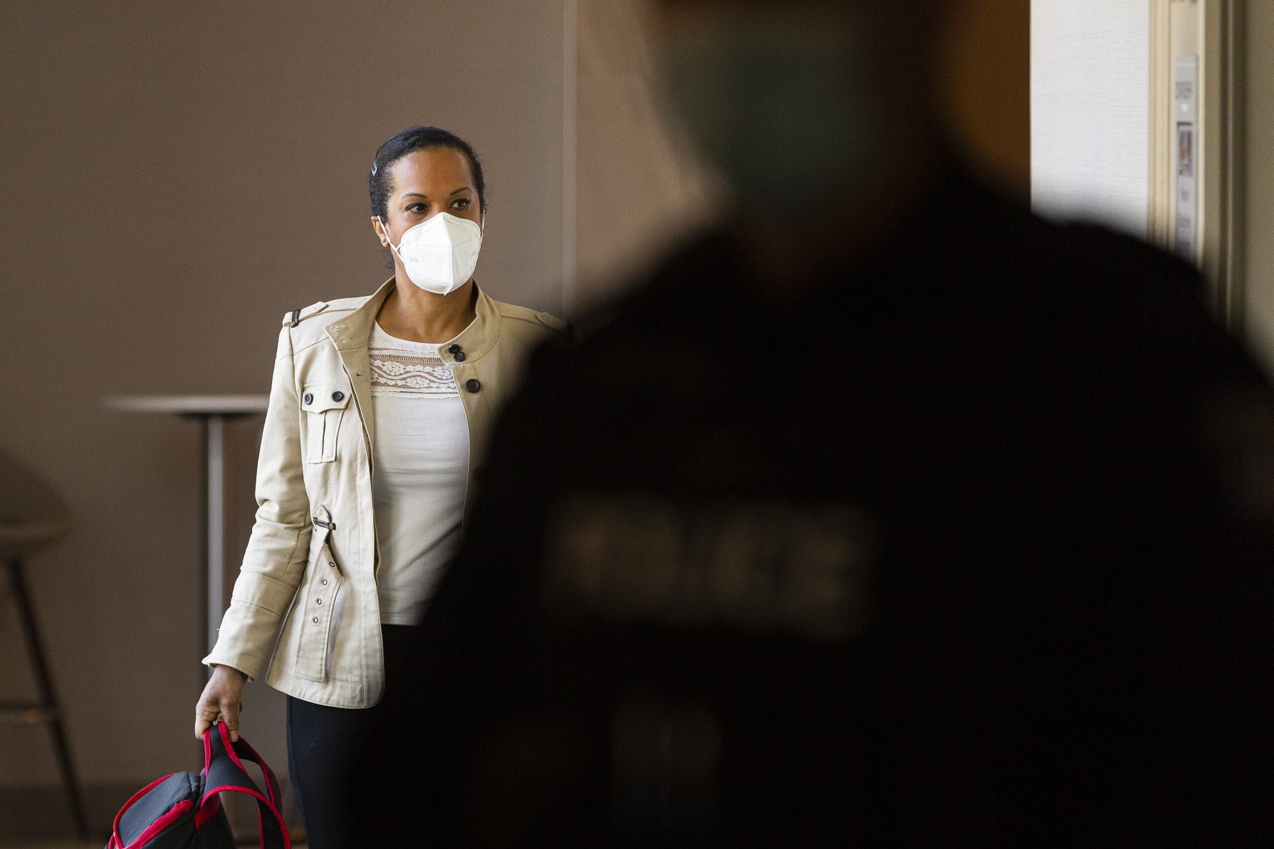 穿着白色面具和棕色夹克的女人，拿着背包，走在旅馆走廊，向她留下来。在前景中，一名模糊的警察穿着面具穿过现场。