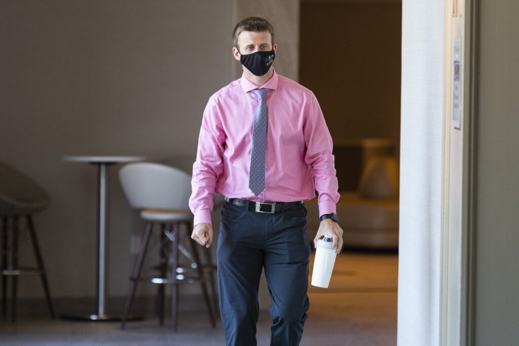 一名男子身穿粉色衬衫，打着灰色领带，戴着黑色面具，左手拿着一个白色水瓶，右手握紧拳头，走在酒店的走廊上。