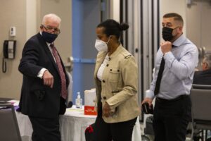 穿着黑暗面具的衣服的一个男人，一名棕色夹克的妇女穿着一件白色面具，和一个男人在衬衫和黑色领带和黑色面具上被看见在会议室。