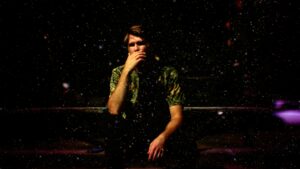 亚当·沃伦的照片。他穿着一件有图案的绿色衬衫，沉思地坐着，用手掩着嘴，在一个非常黑暗和忧郁的房间里，空气中飘浮着一些斑点。