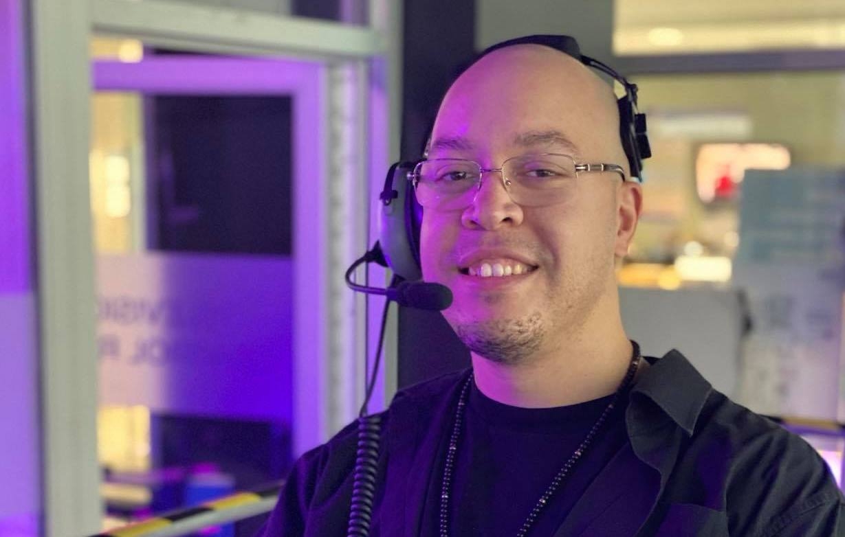 Matthew Byard, in a recording studio. He's smiling at the camera, wearing a headset. He's got wire-framed glasses, a shaved head and the beginnings of a small beard on his chin.