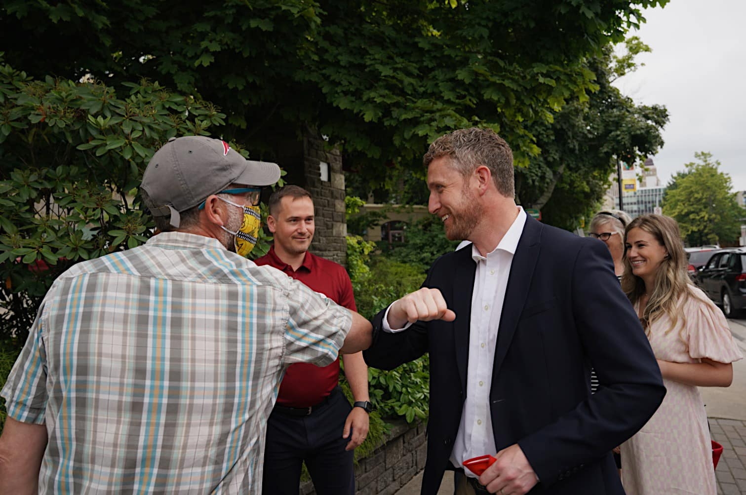 Ian Rankin, with his red mask in his hand, bumps elbows with a man who is wearing a mask. Three other people, maskless, stand close by. One is Rankin's wife.