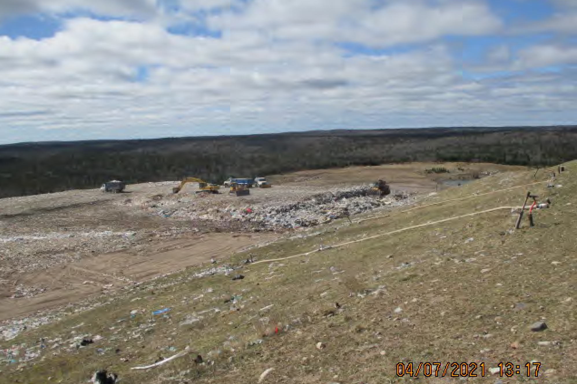在一张时间戳为2021年4月7日13时17分的模糊照片中，远处的卡车和装载机正在填埋地压缩垃圾。这是一个阳光明媚的日子，有些云彩，垃圾填埋场后面有一片森林。