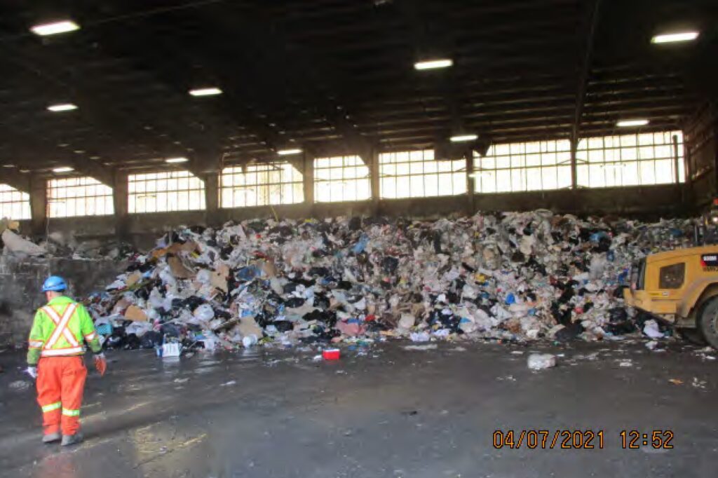 在这张纹理模糊的照片中，时间是2021年4月7日12点52分，一大堆垃圾堆积在一个仓库式的房间里，天花板上有灯，背景中的窗户横跨墙壁。在左边，一名身穿显眼的蓝色、荧光黄色和橙色衣服的工人朝垃圾走去。在右侧可以看到前端装载机的后端。