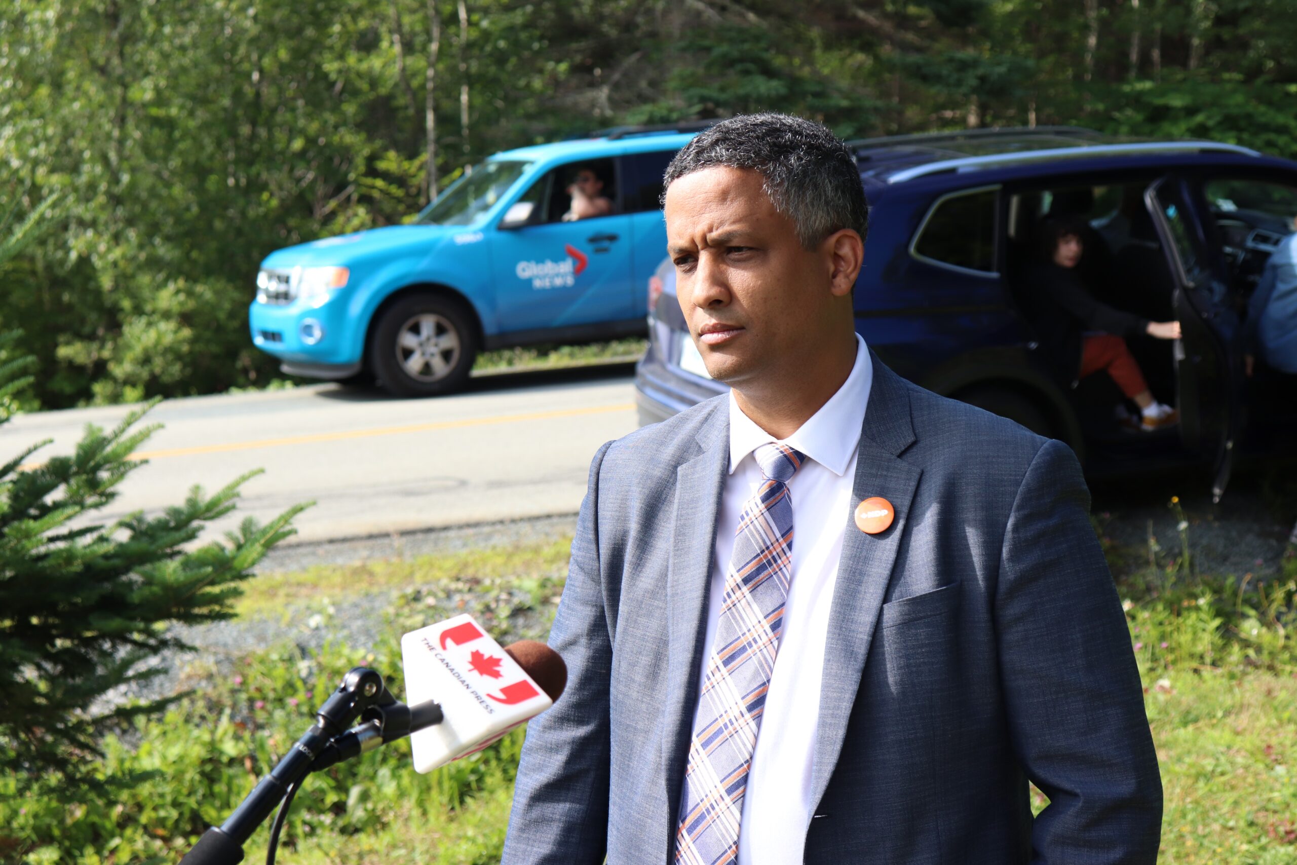 一名身穿西装、戴着新民主党别针的黑人男子在草地上与记者交谈，背景是道路上的车辆。