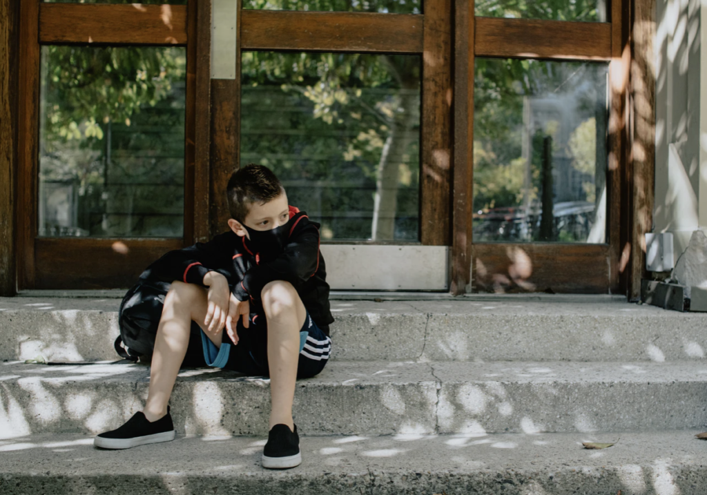 戴着面具的一个幼儿坐在学校台阶与他旁边的背包。