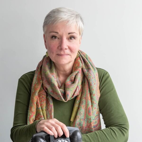 穿着绿色长袖衬衫，系着围巾，身穿白色短发，相貌出众的女子站在一个中性的背景前。