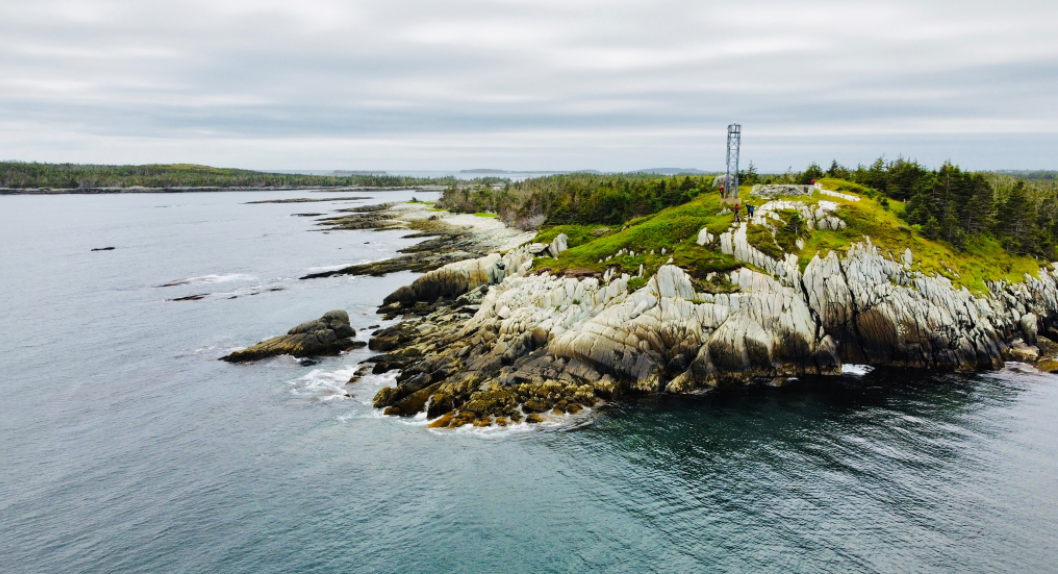 A photo of a crop of rocky land jutting out to the coast. A few people are standing atop the rocky area where there is also a tower.