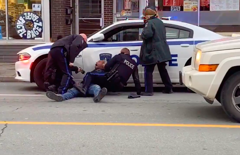 在一段视频的截屏中，四名警察包围了一名躺在地上的黑人，他被压在一辆贴着蓝色贴纸、亮着警灯的白色警车上。左边的一名警官拿着泰瑟枪。左边的另一个趴在副驾驶身上。在右边，一名警官将腿压在男子身上，另一名警官在一旁看着。这一场景是从街对面拍摄的。画面右侧有一辆车停了下来。在背景中，“百吉饼”这个词出现在商店橱窗的圆形贴花上。