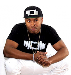 科里·赖特(Corey Wright)自信地看着镜头，戴着一顶黑色棒球帽，身穿印有配套标志的黑色T恤，白色牛仔裤。