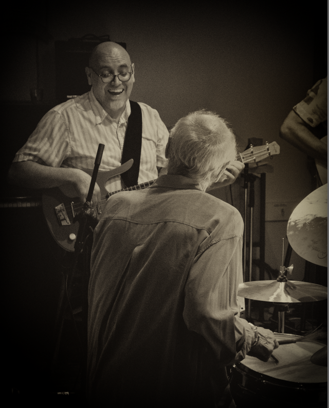 一个男人在弹吉他，面带微笑，看着一个年长的鼓手(我们看到鼓手的后脑勺)