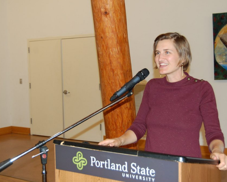 她的30多岁的微笑的妇女站立在指挥台，讲话到麦克风。领奖台有一个横幅，说波特兰州立大学
