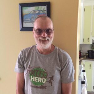 西蒙·斯奈德，在家中对着摄像机微笑。他留着剪短的灰白头发，留着灰白的胡子，戴着一副有色眼镜。