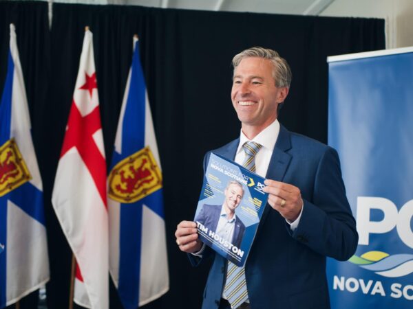 蓝色衣服的微笑的人在Nova Scotia和Mi'qmac旗子前面站立，拿着与他自己的一个文件在盖子。