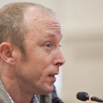 秃顶的白人男子道格拉斯·布兰对着麦克风认真地讲话。