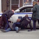 一个黑人坐在街上，背靠着一辆警车，一个警察坐在他身上，另一个警察用泰瑟枪指着他，一个靠在他身上，还有一个站在旁边看着。