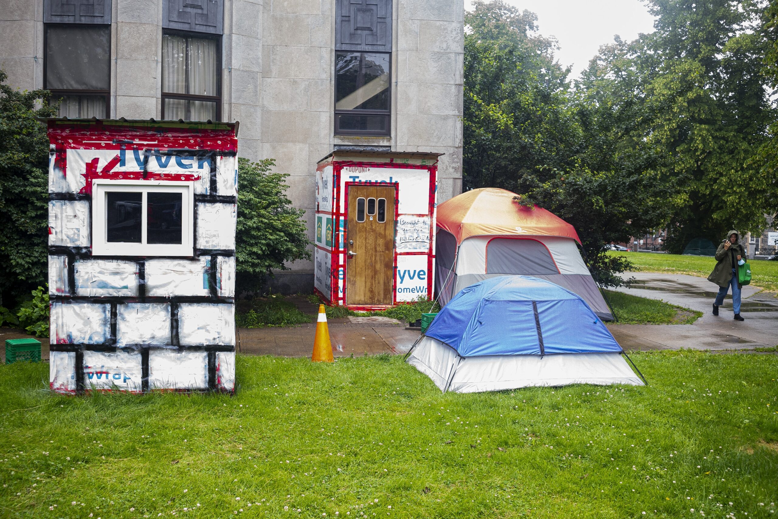 在下雨天，可以看到砂岩建筑外的草地上有两个危机避难所和两个帐篷。左边的遮蔽物是白色的，在可见的一面有一个小窗户，覆盖着涂有黑色网格的特卫强房屋包裹物。在中心，有另一个避难所，有一个木门和三个小窗户。在那个避难所旁边，有一个红灰相间的帐篷。在红灰色帐篷的前面有一个蓝灰色的帐篷。在右边，一位身穿四分之三长外套、提着绿色可重复使用的购物袋的女士一边走一边用免提打电话。