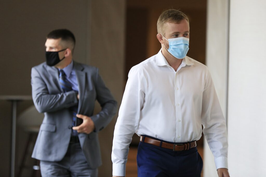 两个人走在旅馆的走廊上。右边的人戴着蓝色的面具，穿着白色的衬衫，蓝色的裤子和棕色的腰带。他在看他的左边。左边后面的男子戴着黑色面具，穿着灰色西装、蓝色细条纹衬衫和蓝色黑色领带。他在看他的右边。