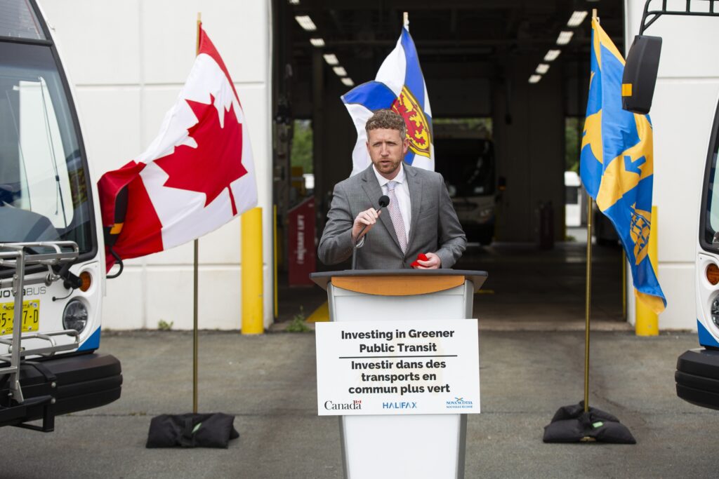 一个男人穿着灰色西装，白色衬衫和虚线在讲台上讲话。在讲台上，它说，“投资更环保的公共交通”，徽标代表加拿大政府，哈利法克斯和新斯科舍。