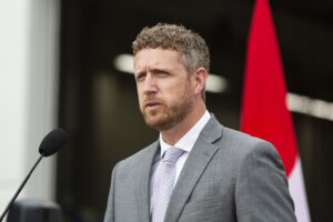 一位身穿灰色西装、白色衬衫、打着圆点领带的男士在讲台上发言。在他身后，可以看到加拿大国旗的一部分。