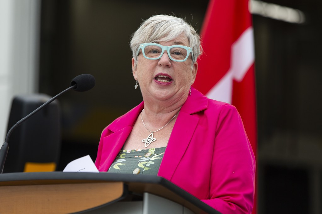 A woman wearing a pink blazer and teal glasses speaks at a podium. Behind her is a Canadian flag.