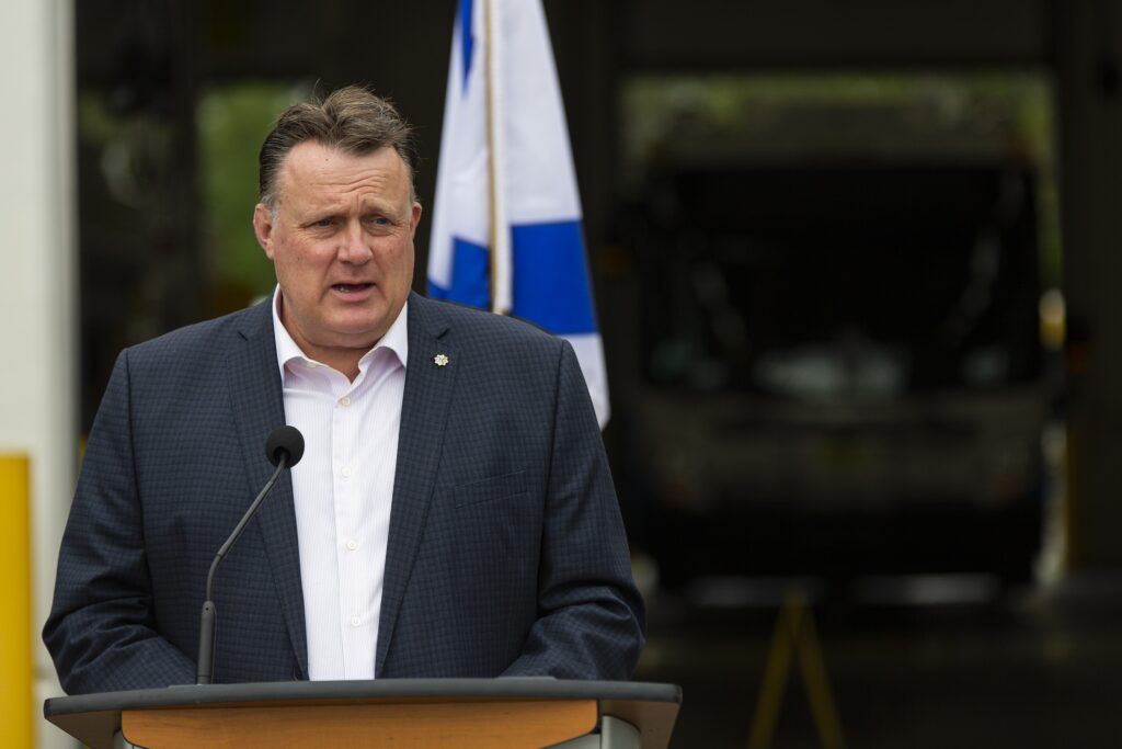 一名穿着黑暗的西装和白色衬衫的男人在讲台和姿态讲话。在背景中是新星斯科蒂亚国旗，蓝色和白色的一部分，以及黑暗中的公共汽车。