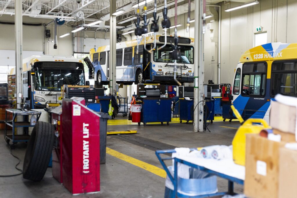 在一个大型车库中看到了三个白色，蓝色和黄色公共汽车。一辆公共汽车在电梯上。在前景中，有一个轮胎靠在标有“旋转升力”的红色盒子上。