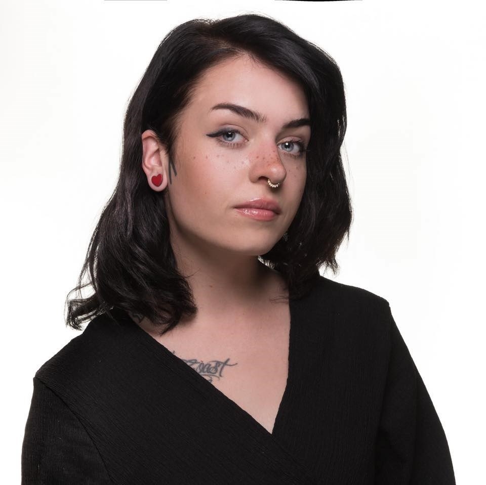 一名女性的头像，棕色长发及肩，v领上衣，右肩下方有部分纹身。