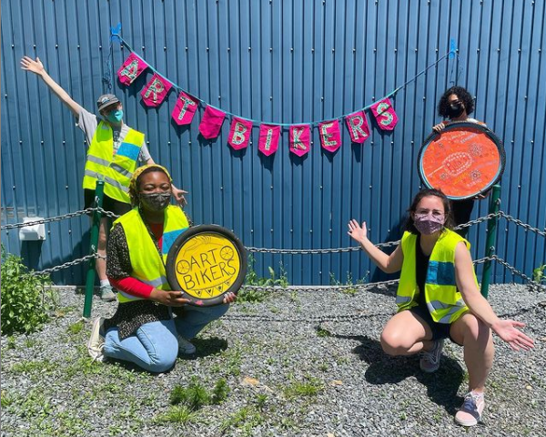 在蓝色墙壁前面的四个被掩没的年轻人。其中两个是站立，两个是跪着的。他们看起来快乐。墙上的横幅和一个人的迹象表明艺术骑自行车的人。