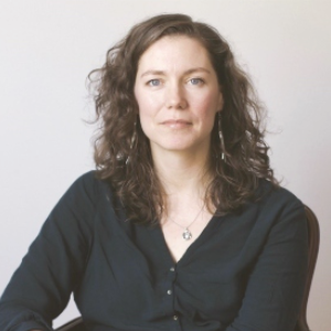 凯瑟琳麦克兰直接看在相机。她有肩长的波浪棕色头发，戴着一件黑色衬衫。