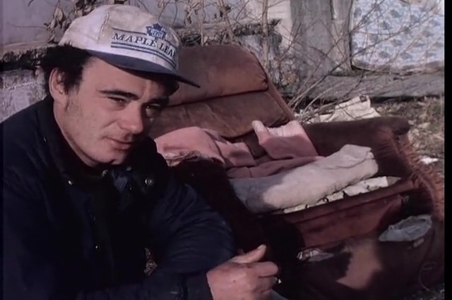 有一个有些麻烦的表达的年轻人，戴多伦多枫叶帽子。他坐在一个被遗弃的地段里，他周围的碎片或垃圾，包括破碎的家具。