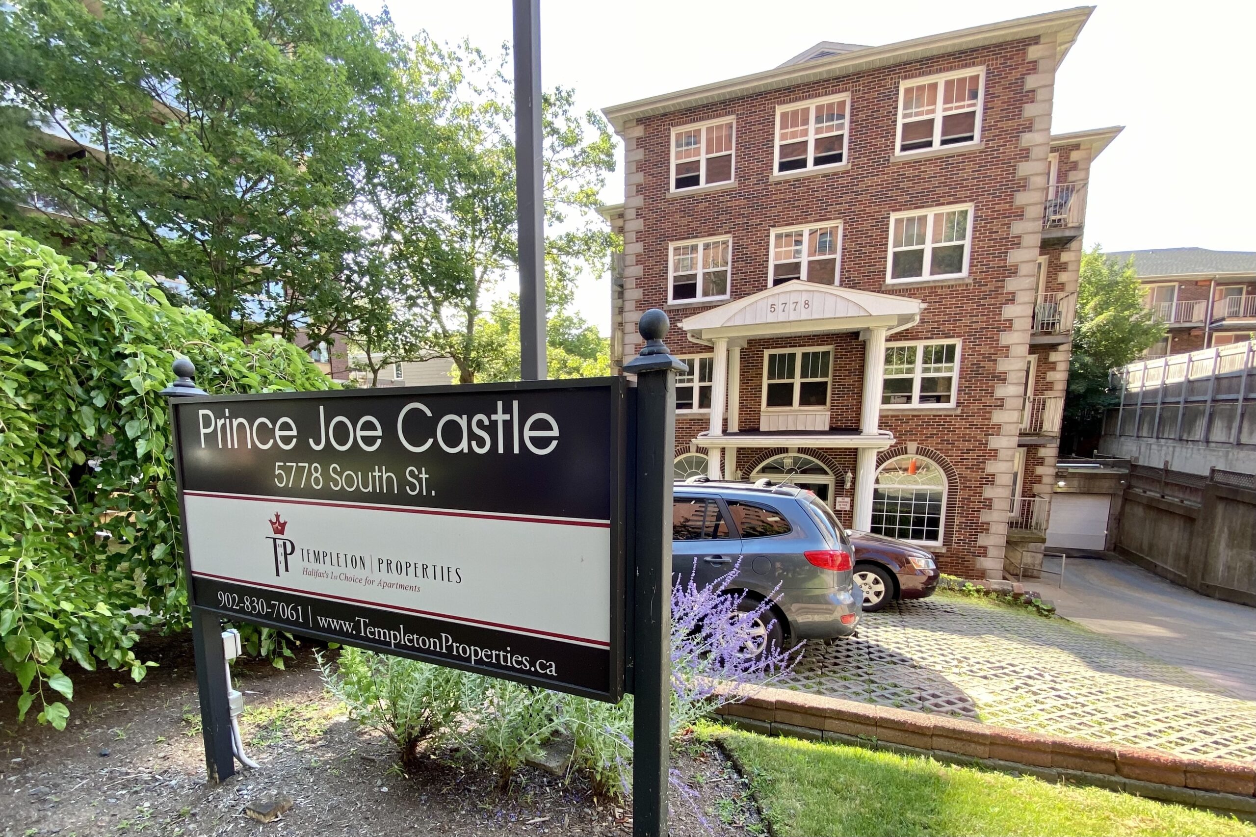 一个四层的砖砌建筑，前面有一个黑白的标志。牌子上写着:“乔王子城堡，南街5778号，坦普尔顿物业，902-830-7061,www.TempletonProperties.ca。”在标识和建筑之间的空地上停着两辆车，路面铺着交错的砖块。左边是绿色的树木和灌木。