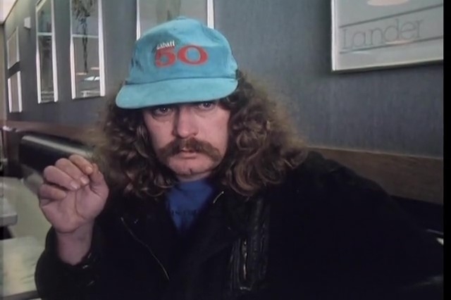 一个留着长长的卷发和下垂的小胡子的男人坐在地板上，戴着一顶Labatt 50的帽子。