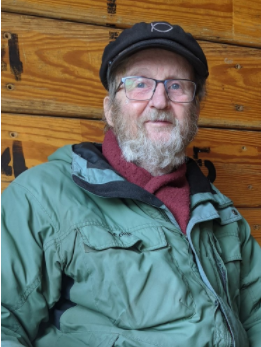 一位留着白胡子、戴着眼镜、身穿防雨夹克、头戴雨帽的老人站在一堵木板墙前。