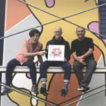 Three people sit smiling on scaffolding in front of a fresh mural.