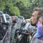 Two male protesters, one wearing a mask, are shown facing a line of police in full riot gear