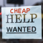 The word cheap is written in red marker over help wanted on a paer sign taped to a shop window.