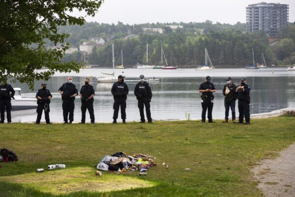 Several cops stand at a waterside park. in the foreground are someone's belongings. And in the background are sailboats in the water.