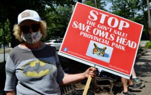 一名戴着帽子、戴着面具、身穿印有蝙蝠侠标志的灰色t恤的妇女在集会上举着一个红色的牌子，上面写着“停止政府销售!”拯救猫头鹰头省立公园