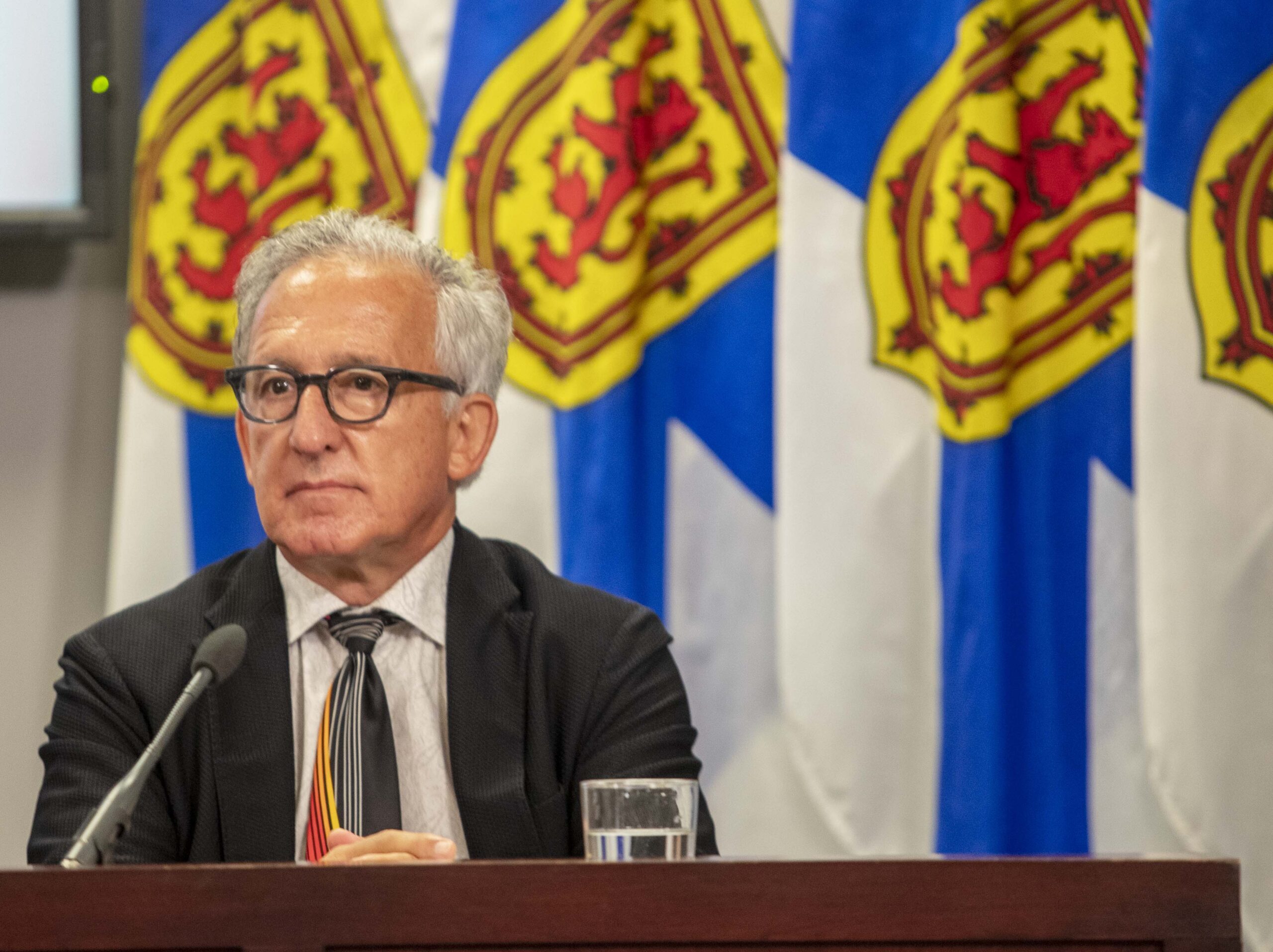 一个白头发、戴眼镜、穿黑色西装的白人坐在桌子旁。他身后是几面新苏格兰国旗