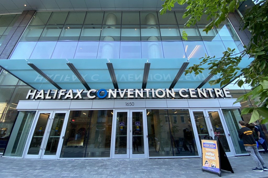 A glass building with a white sign reading, Halifax Convention Centre. The 'O' in convention is blue. In the upper right corner, there are green leaves from a tree hanging into the frame. Below the leaves is a man in a black and yellow jacket waiting beside a yellow sign reading, 