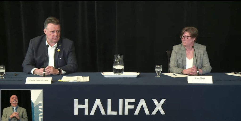 A man wearing a dark suit and a white shirt speaks at a microphone on a table with the HALIFAX logo. To his left is a woman wearing glasses in a grey patterned suit with a white shirt. The woman is looking at the man. Between them is a pitcher of water. The background is black. In the bottom left corner, a sign language interpreter signs.