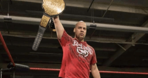 JP·希姆斯穿着一件红色t恤，在摔跤场里举着一条金牌冠军腰带。