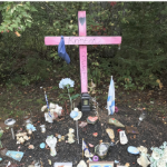 一个粉色十字架的纪念碑，十字架下面有鲜花、旗帜和纪念品