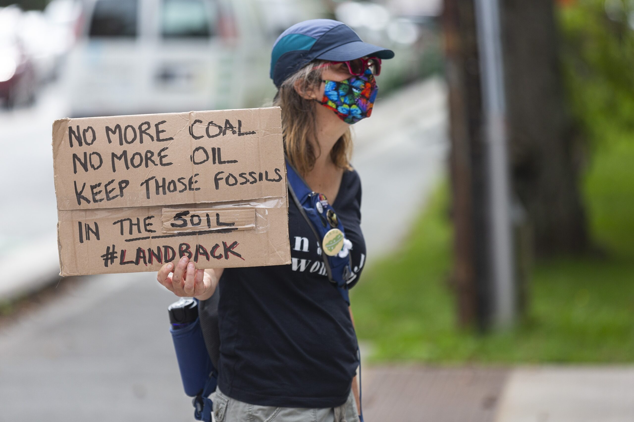 一名戴着蓝色棒球帽和印有花卉图案的面具的抗议者站在纸板上，举着一个牌子，上面写着不要再用煤，不要再用石油，让这些化石留在土壤里#陆地回归#