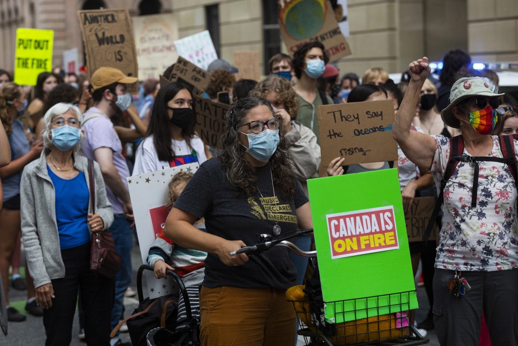 Protesters in a city street, wearing masks and holding signs. One sign, placed in a basket on the front of a bicycle, reads, 