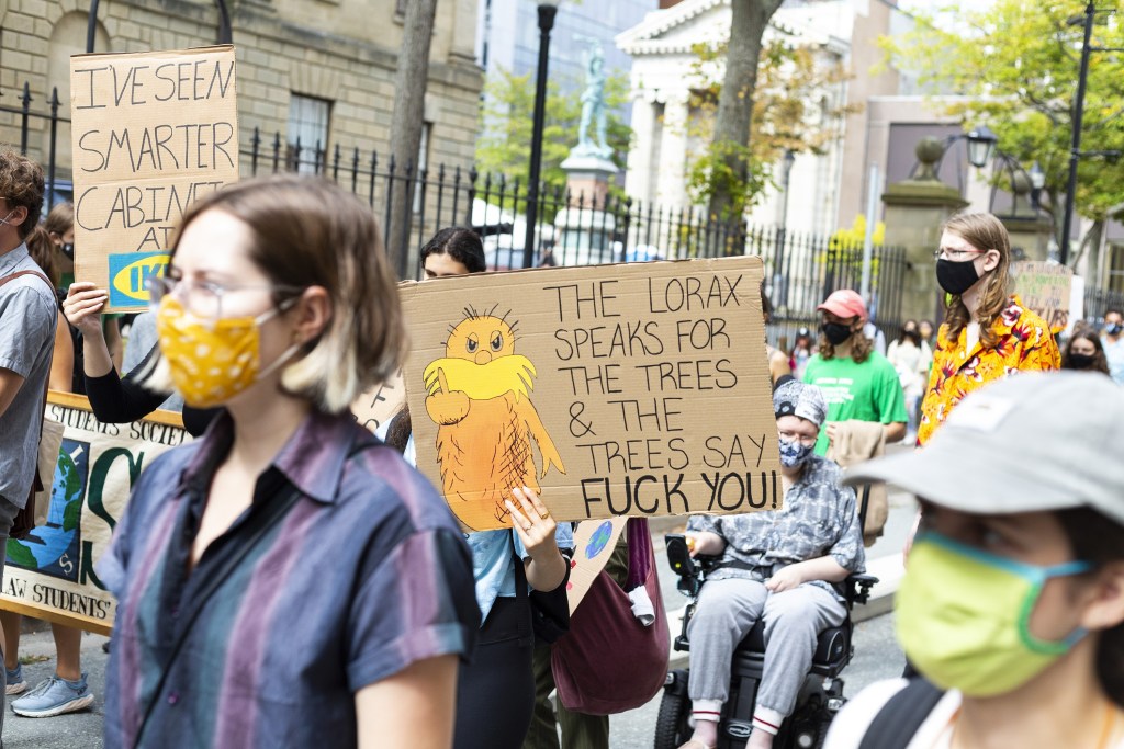 Protesters march through a city street. One holds a sign reading, “The Lorax speaks for the trees & the trees say fuck you!