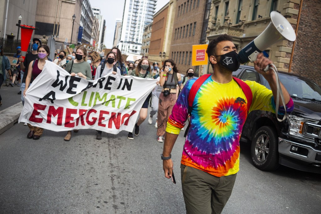 A man wearing a tie dye shirt yells into a megaphone. Behind him, there are hundreds of people marching through a city street hold signs. In the front of the crowd, a few of the protesters hold a banner reading, 