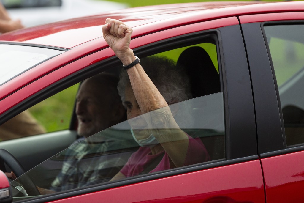 An older woman in a red car holds her fist up out the window. An older man in the passenger seat smiles.