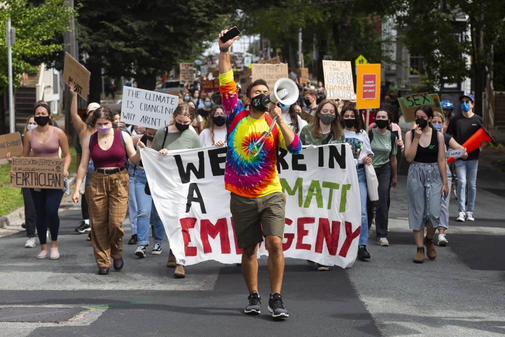 A man wearing a tie dye shirt yells into a megaphone held in his left hand. His right arm is held up holding a phone. Behind, there are hundreds of people marching through a city street hold signs. One say, 